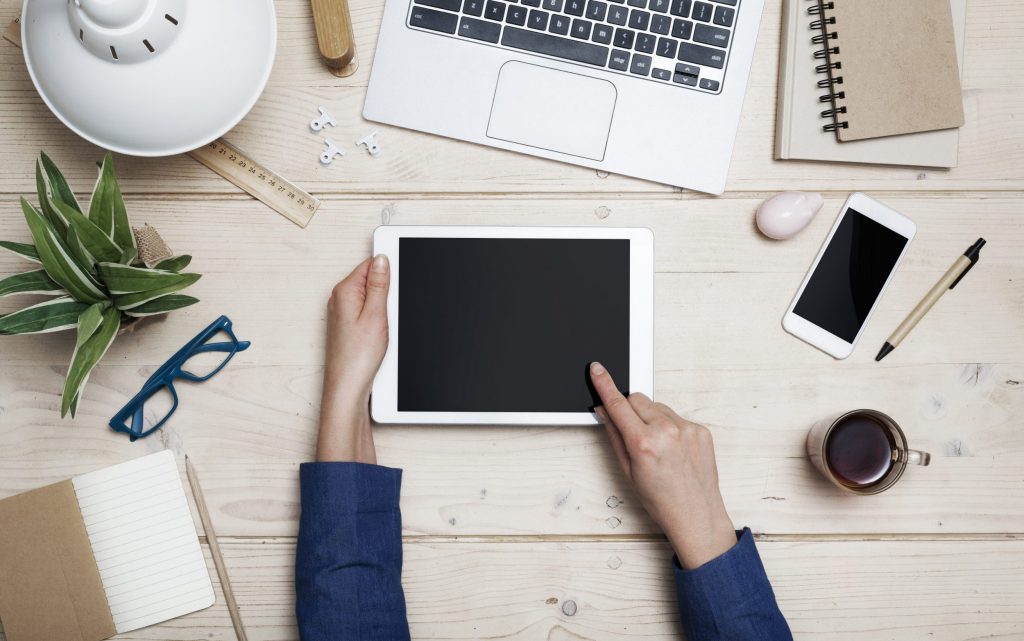 Overhead flat-lay of a wooden desk with hands holding a tablet, surrounded by a laptop, phone, notebooks and a plant, light and tidy mood